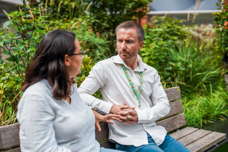 A man wearing a sunflower lanyard to show his less visible disability talks with a friend on a bench.