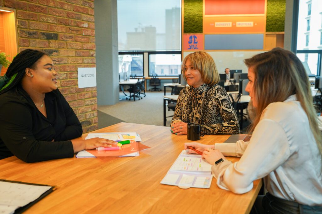 Colleagues with less-visible disabilities meet in the quiet zone of an office, with paperwork, pens and notepads on the table between them. Left, a woman with ADHD, autsim, dyslexia and a mental health condition, middle, a woman with a brain injury and limited mobility, right a woman with vitiligo and diabetes.