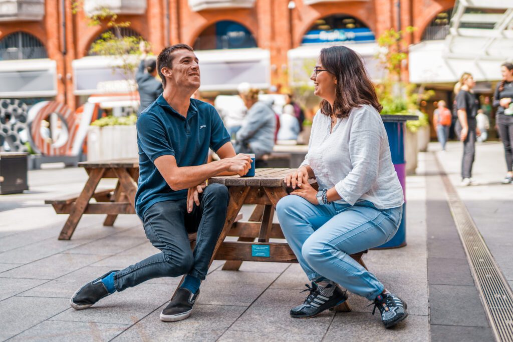 A young man with multiple disabilities sits at a table with a friend at an outdoor shopping centre.