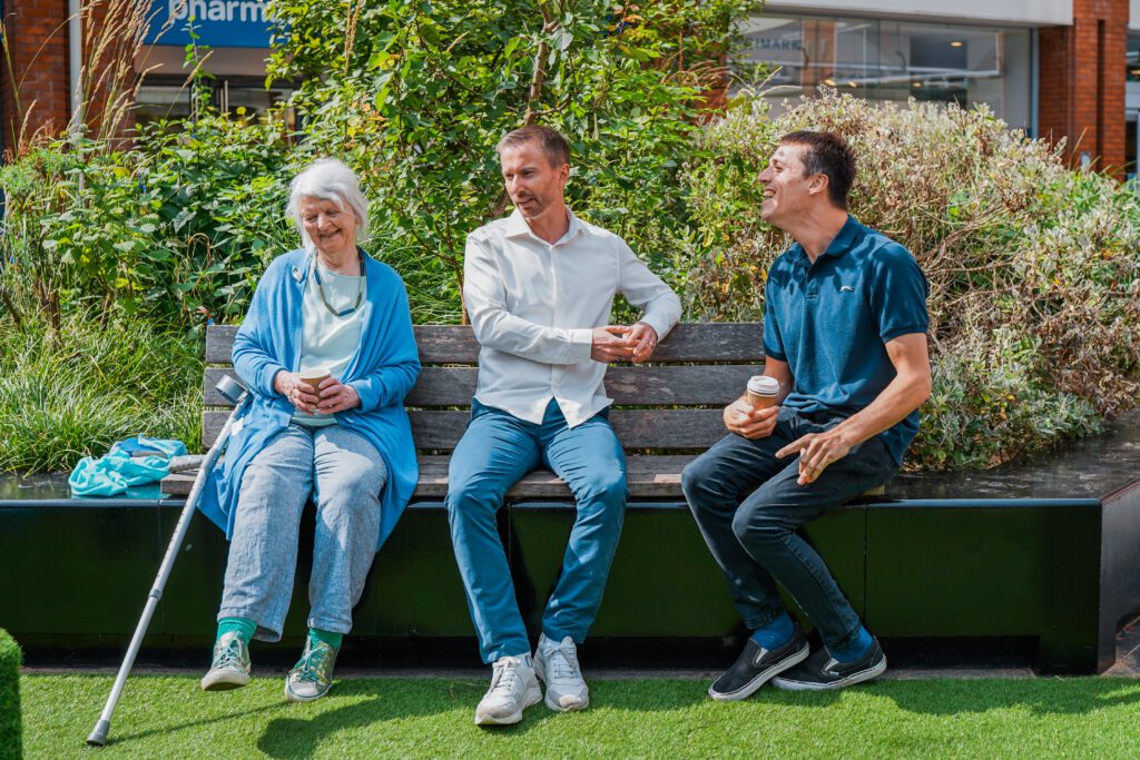 Group of people with diverse disabilities sitting on a bench talking. Left to right: an older woman with a physical impairment who uses a crutch, a man with a less visible disability (early onset Parkinson' Disease) and a young man with multiple disabilities.