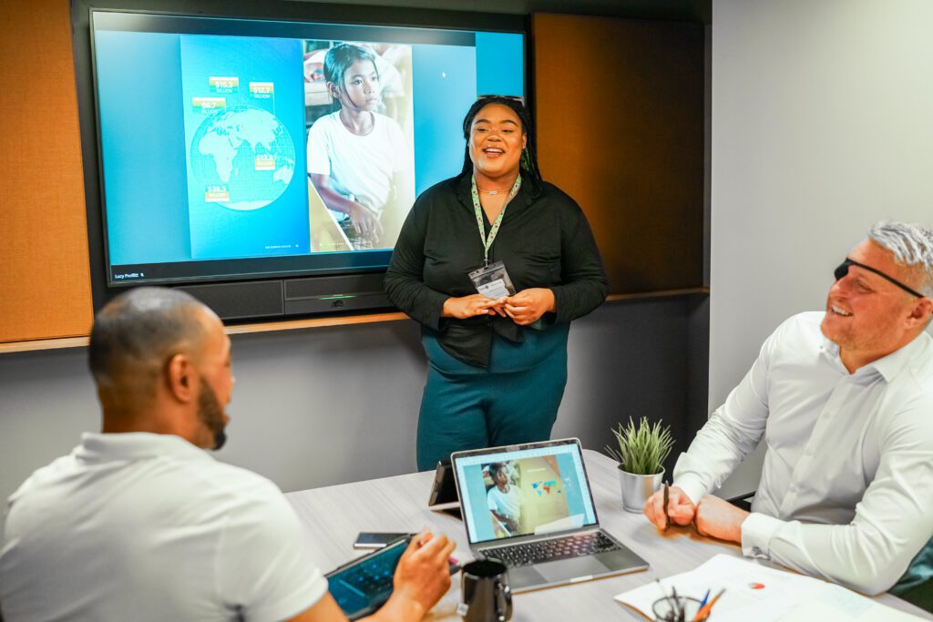 Person with less-visible disabilities delivers presentation to two colleagues, one has a facial difference and sight loss. Left, a man sits at a conference table; Centre, a woman presents with a screen behind her, smiling, Right, a man with a facial difference and sight loss sits at the conference table, smiling.