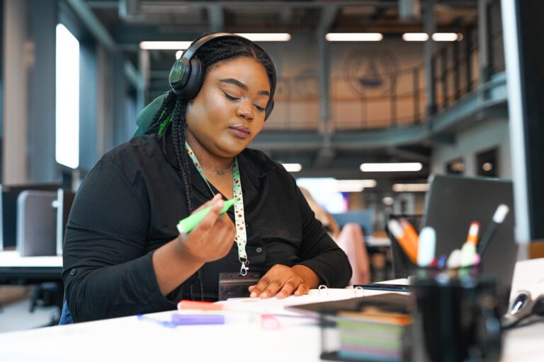 Woman highlights her work at a desk. She is wearing noise cancelling headphones and is wearing a sunflower lanyard to show she has less-visible disabilities.