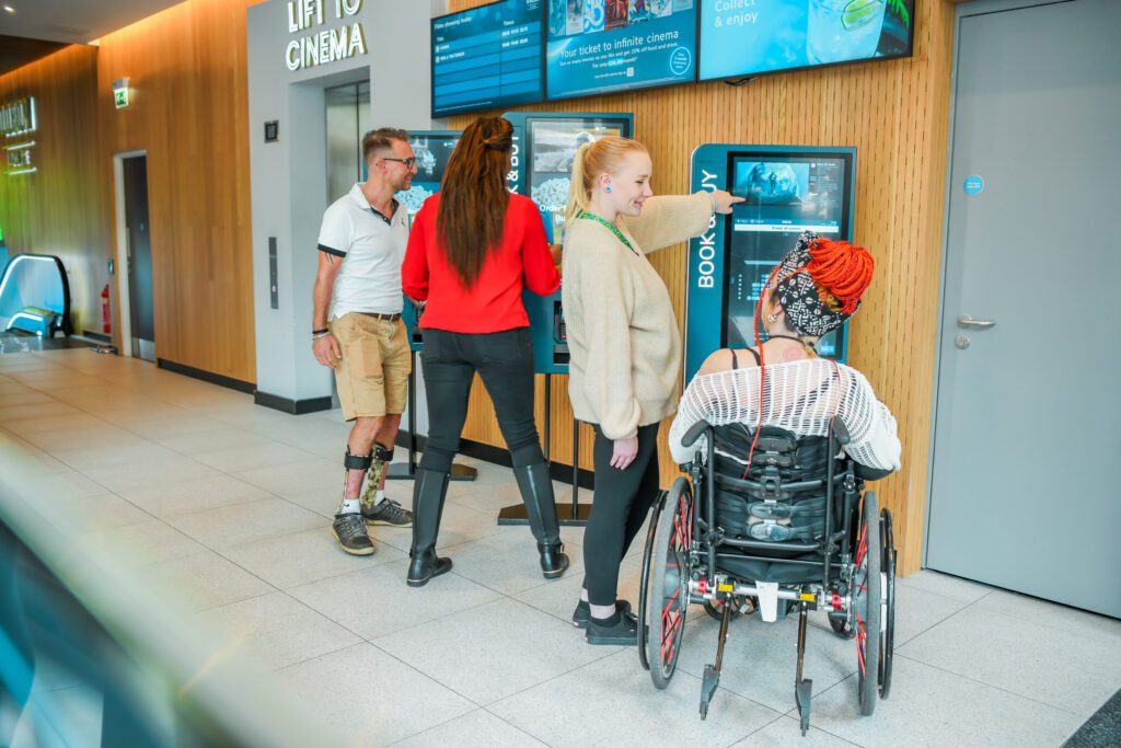 A group of friends make film selection on machine at cinema. Man on the left has splints on his legs and a less visible disability (Diabetes). On the right is a wheelchair user with a spinal cord injury. On her left is a woman wearing a sunflower lanyard to show she has less visible disabilities (multiple).