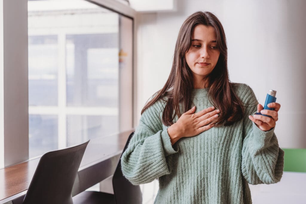 A woman with less-visible disabilities (energy-limiting condition: asthma and Inflammatory Bowel Disease and an Ostomy bag) has one hand on her chest. She holds a blue inhaler in her other hand.