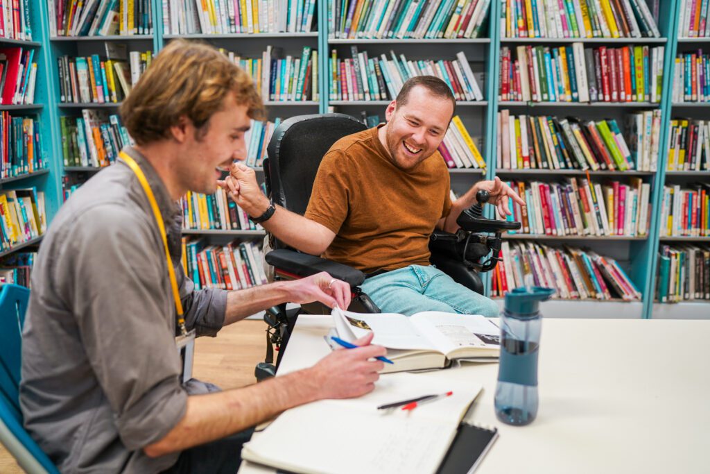 A wheelchair user with Cerebral Palsy works with a friend in the library.