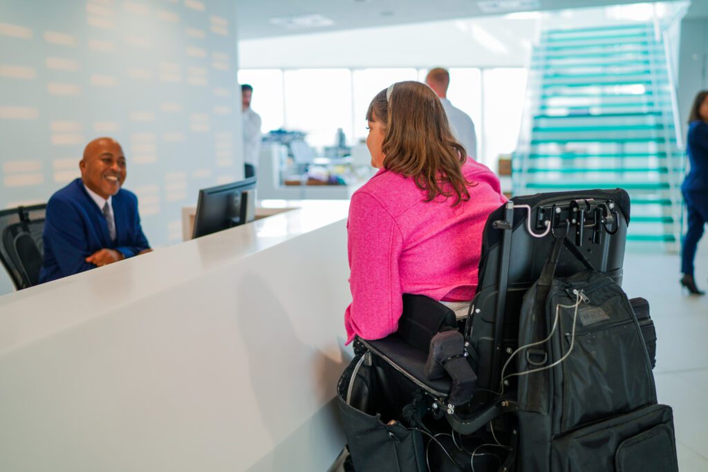 A male office receptionist with a less-visible disability (mental health condition)welcomes a visitor who is a wheelchair user (with Chanarin-Dorfmann Syndrome)