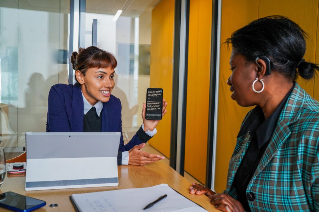 Deaf woman wearing a hearing aid being shown a text message on a smart phone by a colleague with a less-visible disability (mental health condition) in a meeting room.