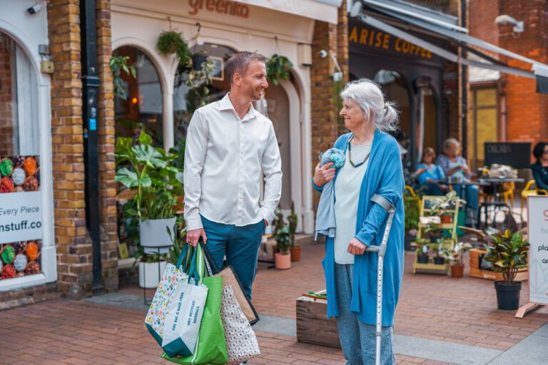 An older woman who uses a crutch shops with a man with a less visible disability (early onset Parkinson's Disease) who is carrying bags.