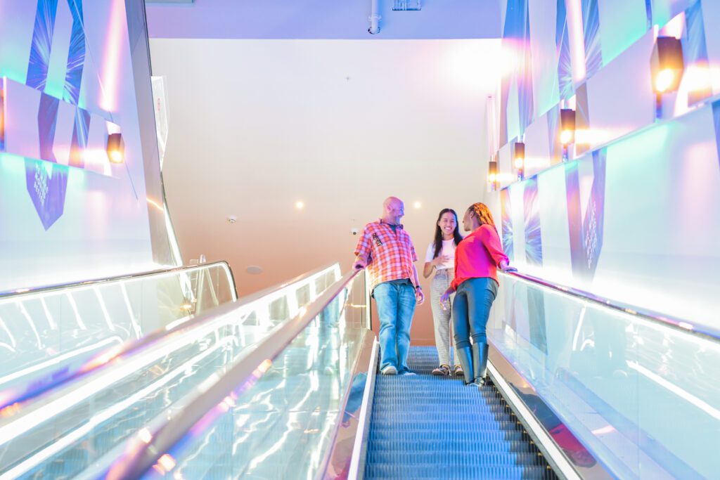 Group of friends laugh at top of escalator at cinema. Man on left has sight loss and a facial difference. Woman in centre is Deaf. Both have long-term health conditions.