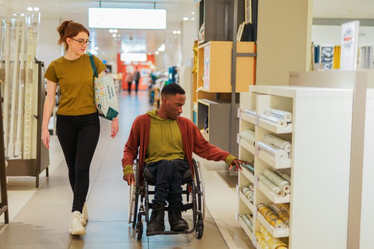 A male wheelchair user with Spina Bifida shops with a friend who has a less-visible disability(PMDD)