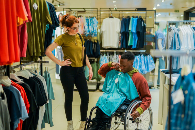 A male wheelchair user holds a T shirt up in a clothing store to show a friend.