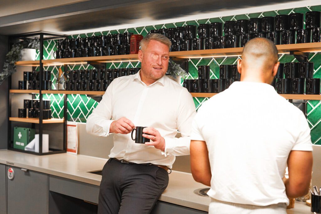 Two men talking in a bar area, one has sight loss and facial difference.