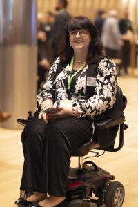 Woman with long black hair and glasses who uses a wheelchair, attending an event