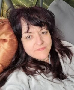 Woman with zero make up and long brunette hair, lying on a brown cushion on garden swing, looking at camera.