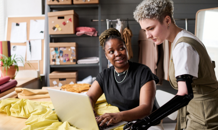 Two women working in a creative studio and looking at a laptop. One of the women has a physical impairment and has a prosthetic limb.