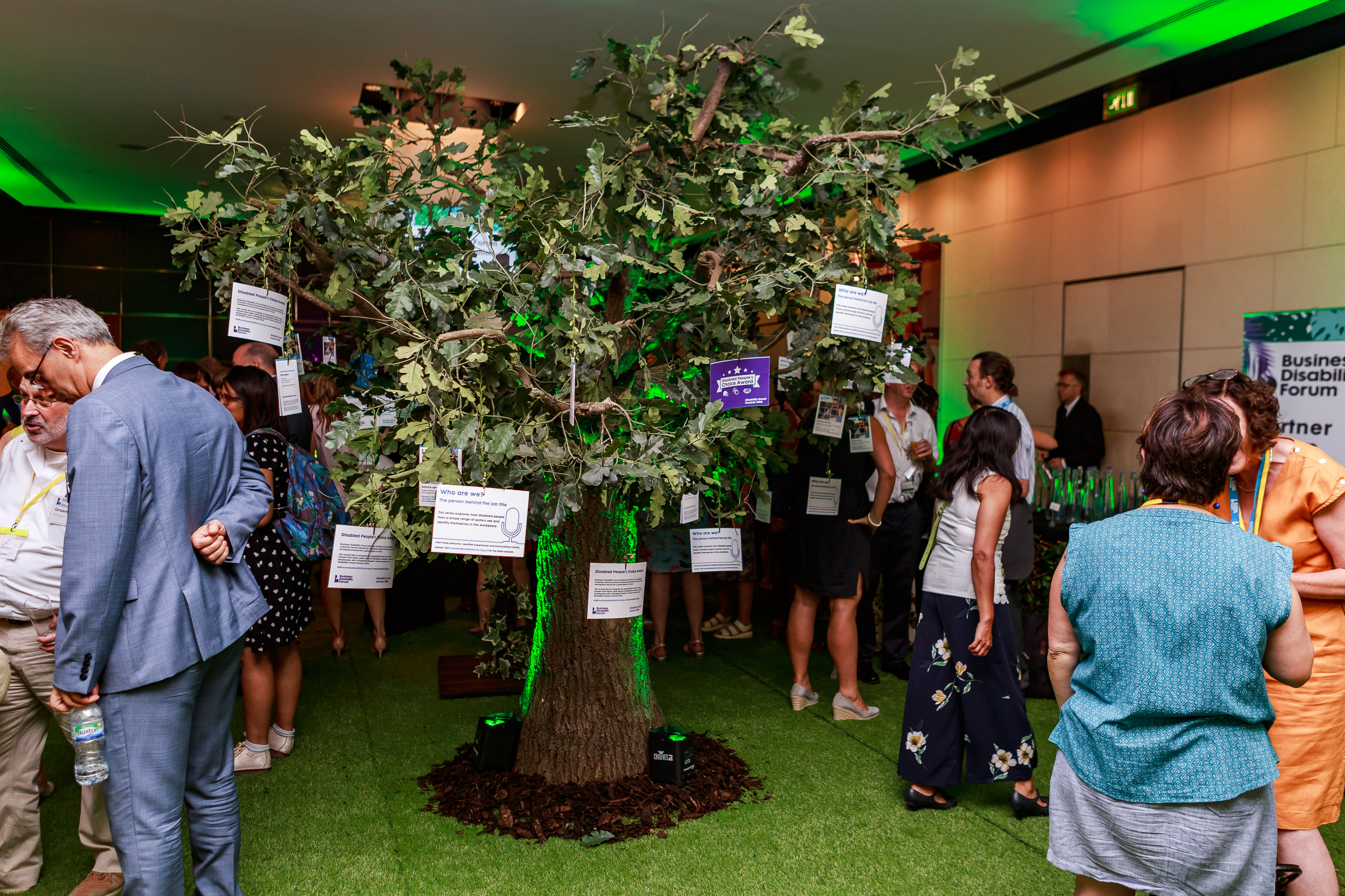 An indoor tree with paper notes hanging from its branches wit people around it at an event.