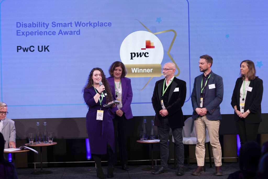 Group of 4 winners on stage at the Disability Smart Awards 2024 ceremony. One of them is holding an award and speaking into a microphone.