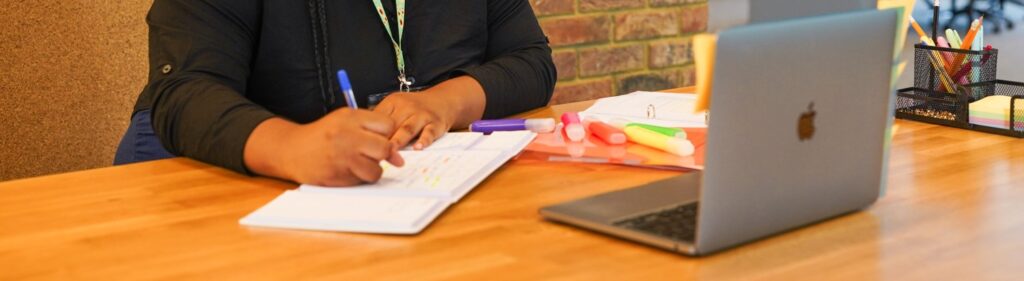 A woman at a desk takes notes on a notepad while a laptop is open across the desk from her.