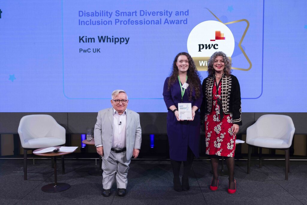 Kim Whippy posing with her award on stage with co-hosts Diane Lightfoot and Simon Minty at the Disability Smart Awards 2024 ceremony.