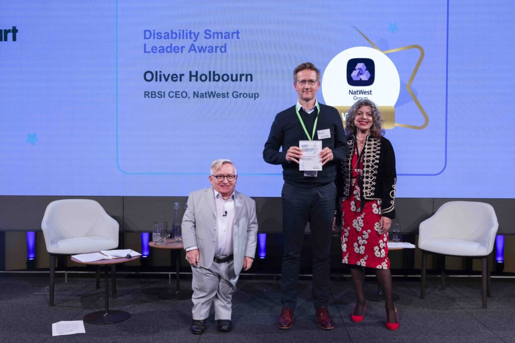 Oliver Holbourn posing with his award on stage with co-hosts Diane Lightfoot and Simon Minty at the Disability Smart Awards 2024 ceremony.