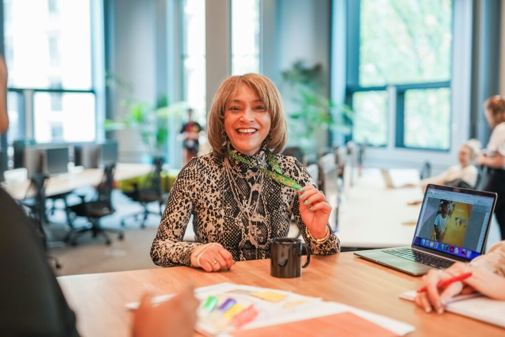 A woman with a less visible disability smiles to colleagues and shows sunflower lanyard in meeting.