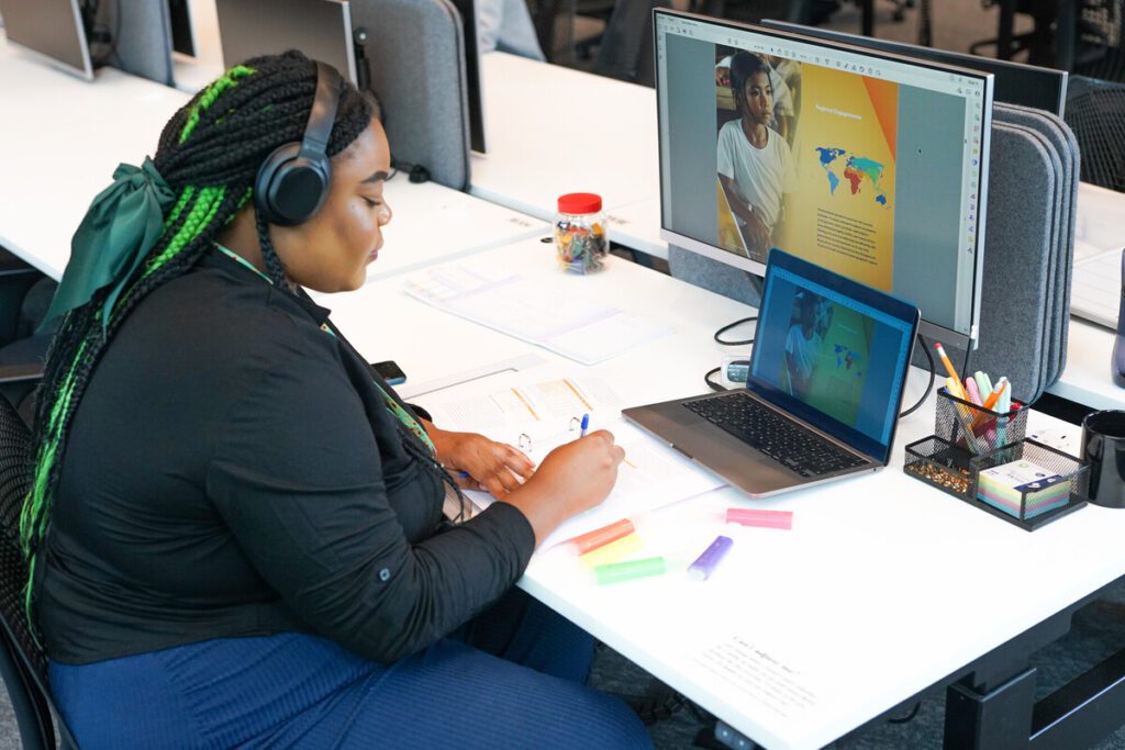 Person wearing headphones in front of a large screen, a laptop and a notebook.