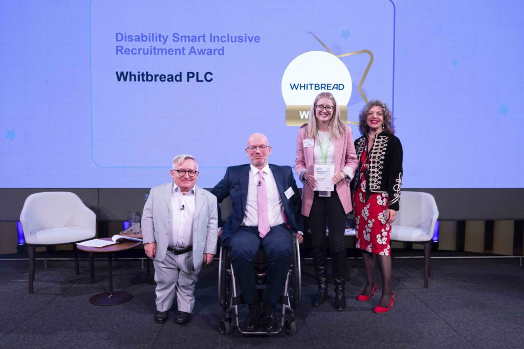 The Whitbread team posing with their award on stage with co-hosts Diane Lightfoot and Simon Minty at the Disability Smart Awards 2024 ceremony.