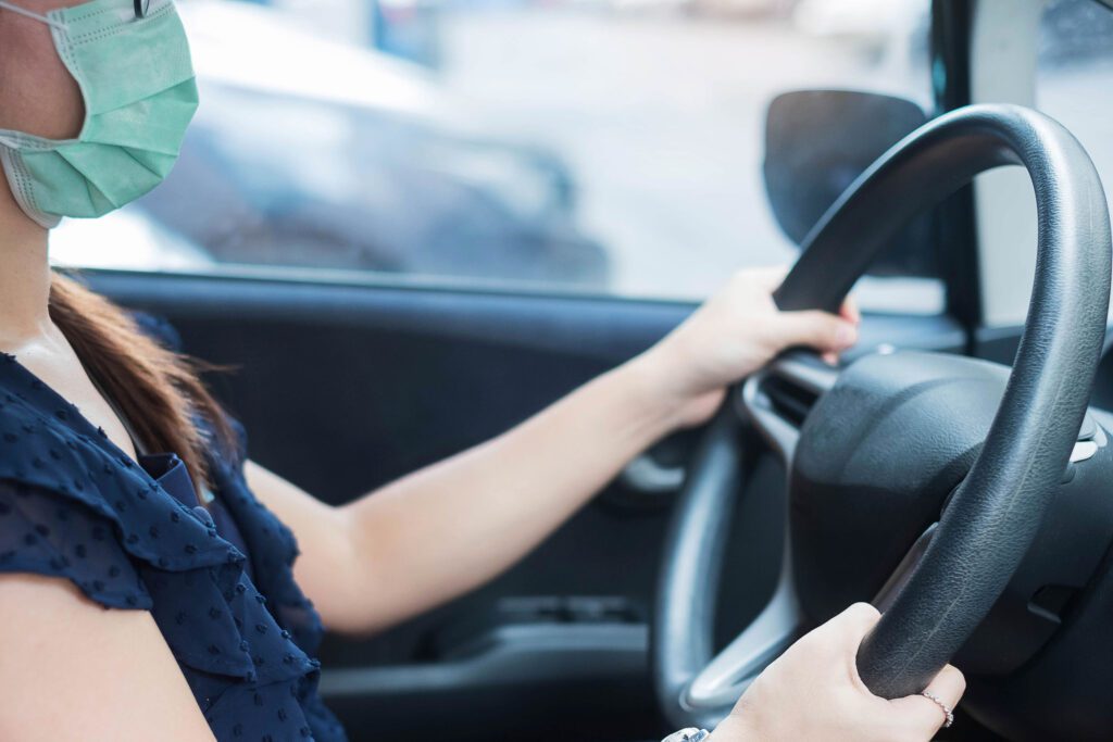 Young woman wearing protective face mask and hand controlling steering wheel in her car