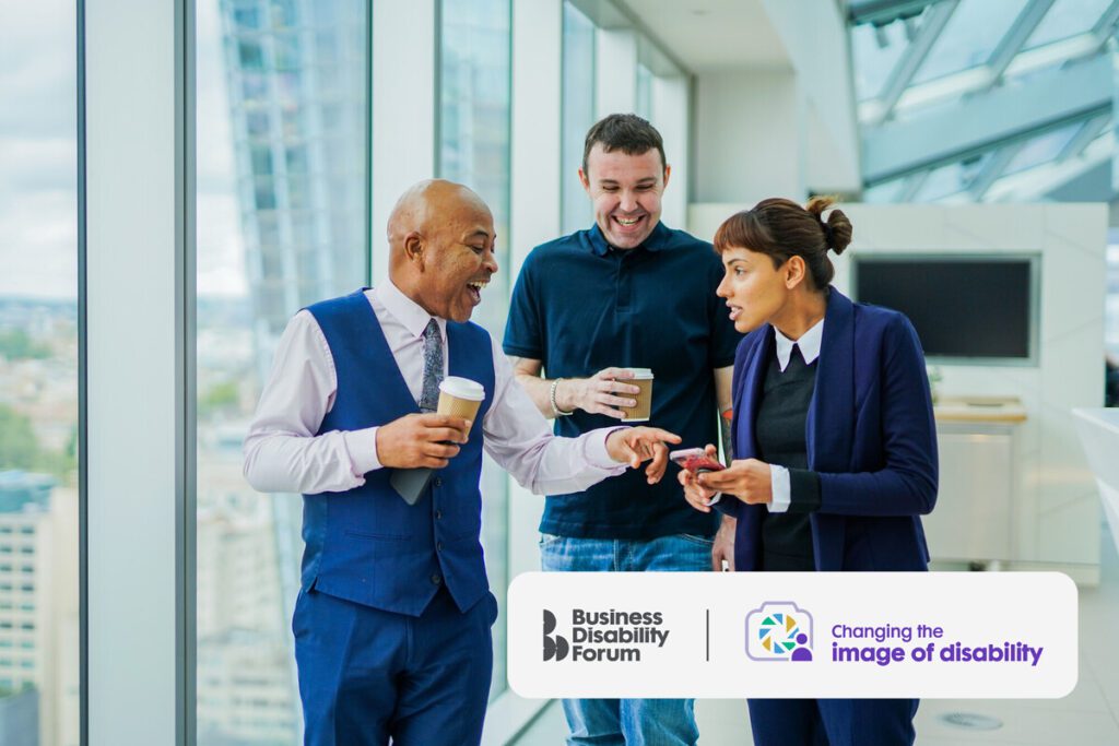 Three colleagues with less visible disabilities walk together laughing and holding coffees in an office.