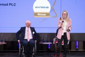 Blonde woman speaking into a microphone on stage at an event. A man in a suit who uses a wheelchair looks on with a smile on his face. The Whitbread logo is projected onto a screen behind them, above a gold box that says 'winner'.