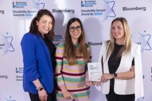 Three women pose with an award. The woman in the middle uses a stick. Behind the group is a banner with the Disability Smart Awards 2024 and Bloomberg logos.