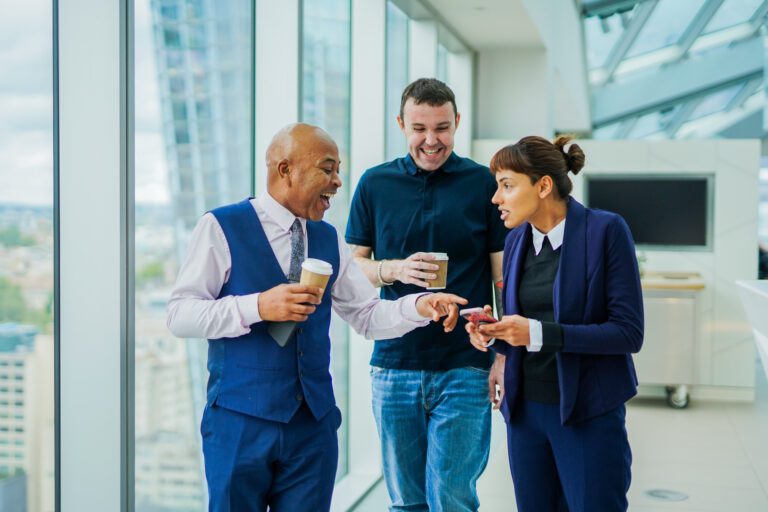 Colleagues with less-visible disabilities walk together smiling with coffees in office. Left, two men holding coffee cups smile and laugh. Right of them, a woman shows them something on her phone screen and speaks.