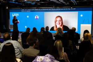 Two speakers addressing an audience at a conference. One is standing on the left of the stage and the other is on video on a big screen. The photo is taken from the back of the audience.