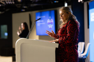 Speaker, who's blind in one eye, giving a speech from behind a lectern onstage at a conference. In the background is a BSL interpreter.