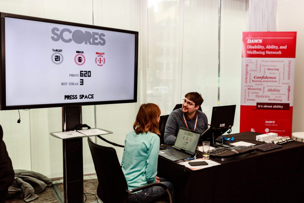 Two people sitting at an exhibition stand at an event, with a big screen behind them.