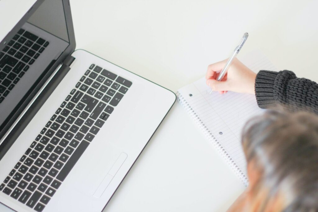 Person writing in a notebook with a laptop open next to them, photographed from ablove.