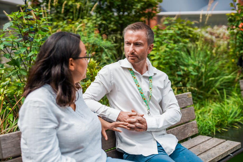 Person with a less-visible disability wearing a sunflower lanyard talks with a friend on a bench .