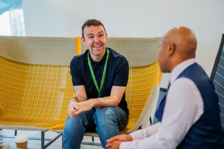 Smiling person with a less-visible disability wearing sunflower lanyard interacts with colleague in office seating area