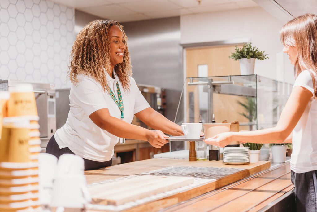 Server wearing a sunflower lanyard (to show they have a less-visible disability) hands tray to customer in a cafeteria.