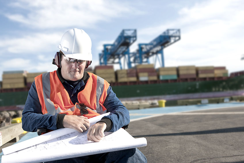 Wheelchair user wearing a safety helmet and a high vis jacket consults plans on a construction site.