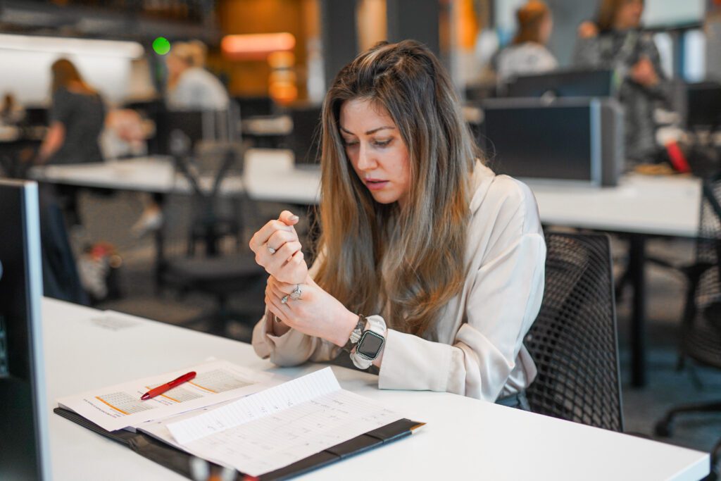 A woman with a less-visible disability sits working at a desk in an open plan office. She is holding her painful wrist.