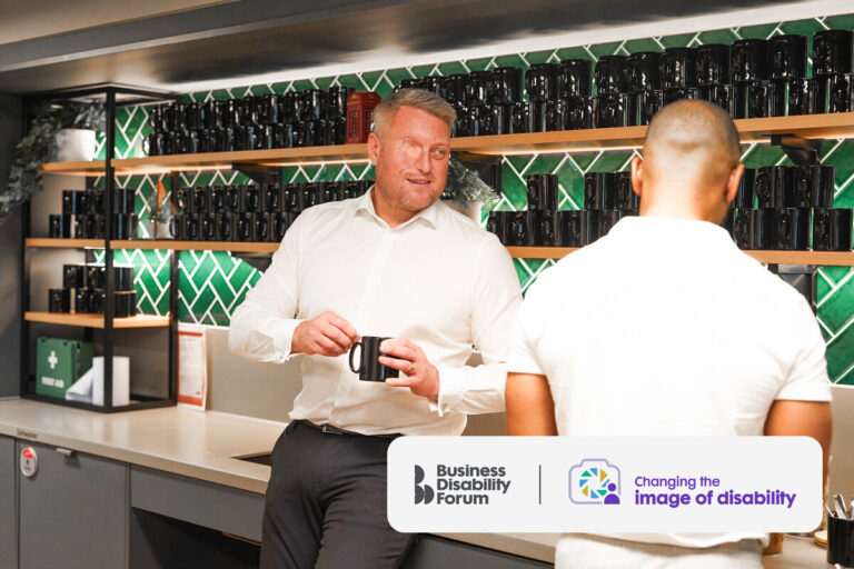 Two colleagues chat while making drinks in a work kitchen area. One has a facial difference and sight loss.