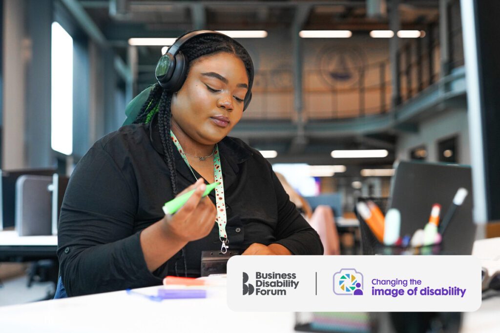 A woman with multiple less-visible disabilities works at a desk. She wears sound cancelling headphones and a sunflower lanyard.