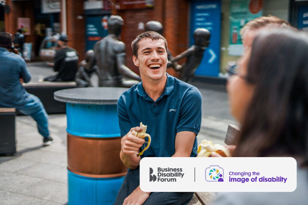 A man with cerebral palsy sits laughing with friends in an outdoor shopping area. They are all eating bananas.