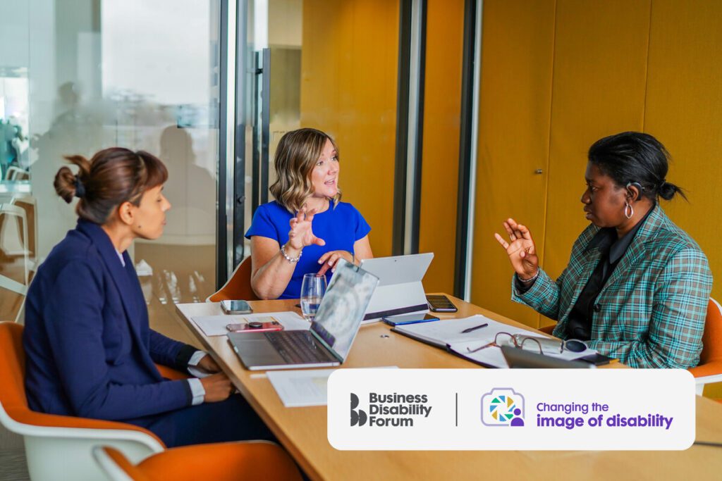 Three seated colleagues talk in a work meeting room. Two have less visible disabilities, one is Deaf and wears hearing aids.