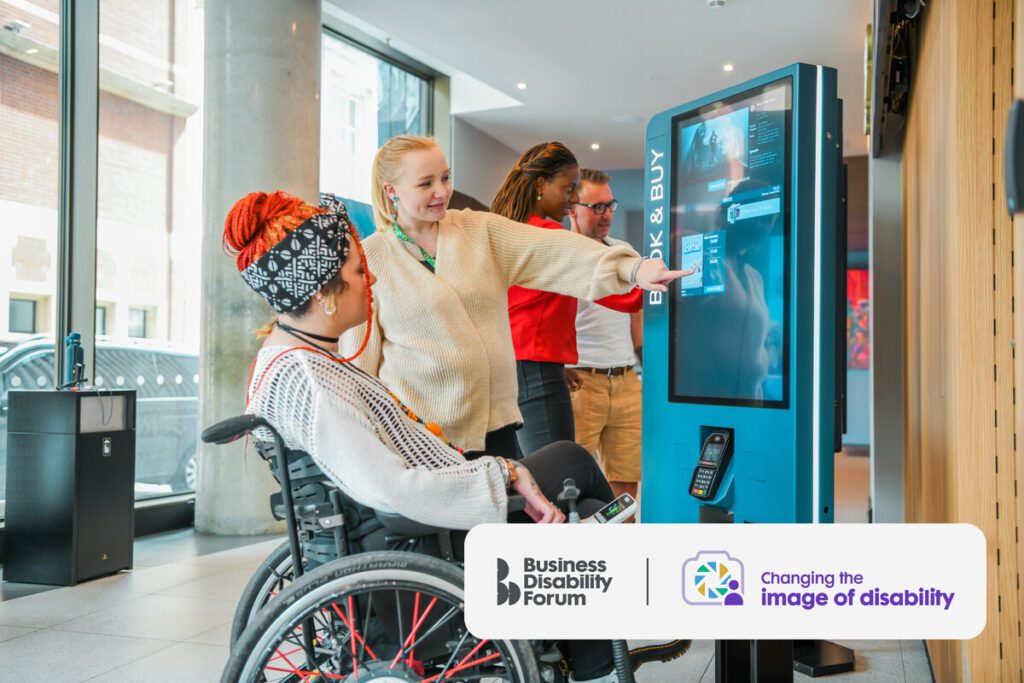 Two friends buy cinema tickets from a touch screen machine. One has a less visible-disability and wears a sunflower lanyard. The other uses a wheelchair.