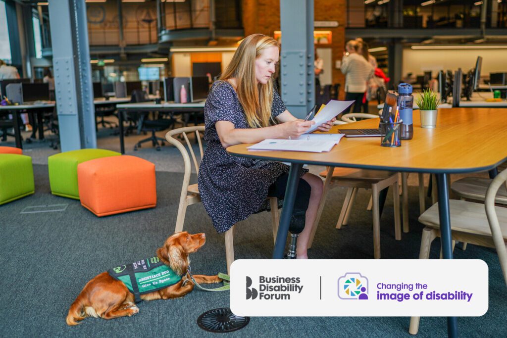 A woman with a prosthetic leg and who has a less visible disability works at an informal office desk. Her assistance dog lies beside her on the floor.