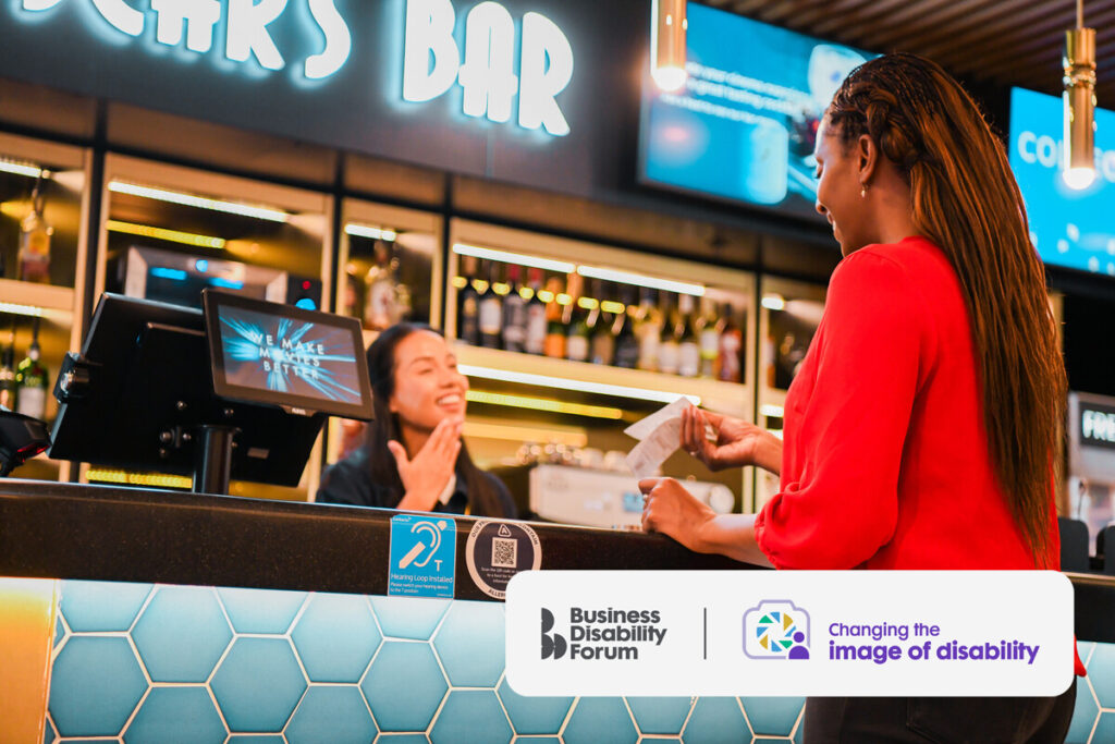 A Deaf sales assistant at the till in a bar signs to a customer while passing over a receipt.