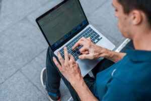 A young man with multiple disabilities uses a laptop while he sits outside. His hands show a physical impairment. 