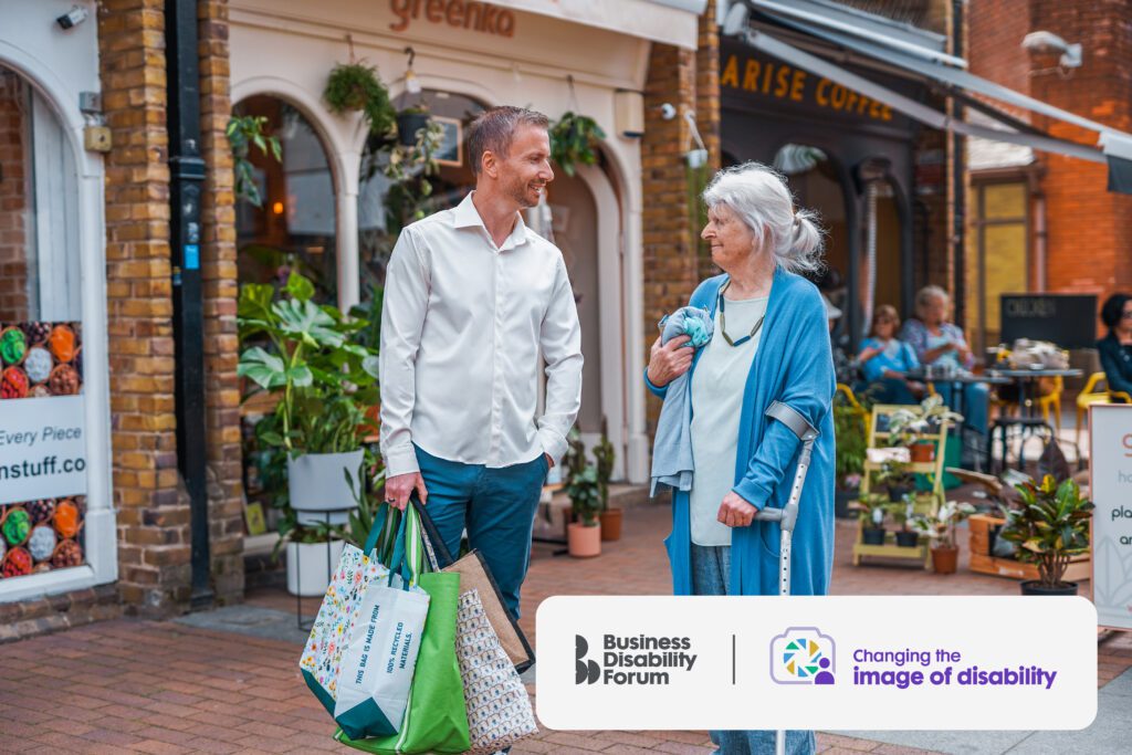 A man with a less-visible disability carries shopping bags and talks to a woman who uses a crutch. They are outside some shops.